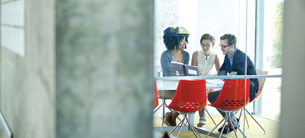 Small team meeting in glass conference room with red chairs, coworkers collaborating on laptop in modern office setting