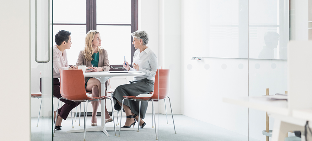 Three professionals meeting at round table in bright modern office, discussing documents and collaborating in glass-walled workspace