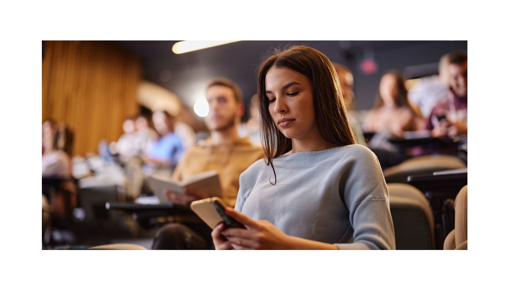 Student using smartphone in lecture hall during class, college classroom audience seated for academic presentation