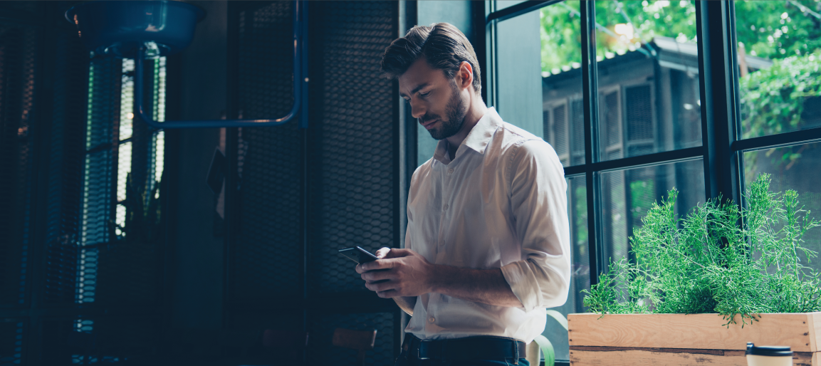 Businessman using smartphone in modern office with large windows and indoor plants, checking messages in bright natural light workspace