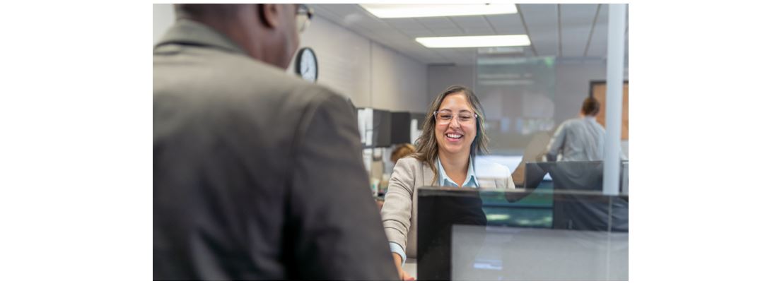 Customer service representative assisting client at office reception desk behind glass, front desk support and visitor check-in