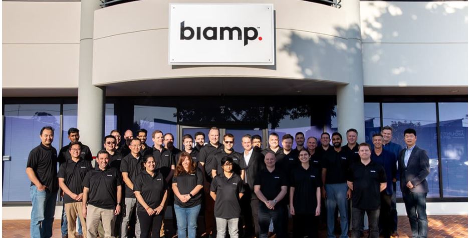 Biamp team group photo outside company headquarters building, employees standing under Biamp sign at corporate office entrance.