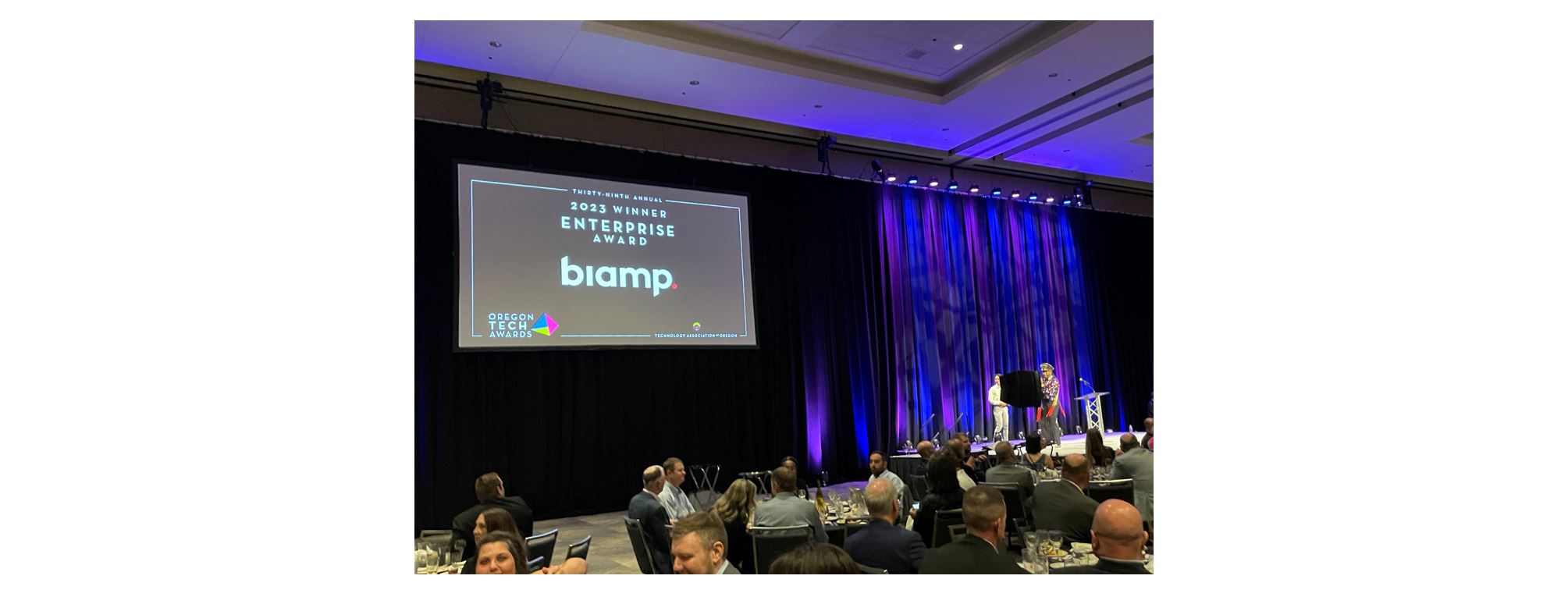 Biamp receiving 2023 Oregon Tech Awards Enterprise Award on stage at banquet event with large presentation screen and audience