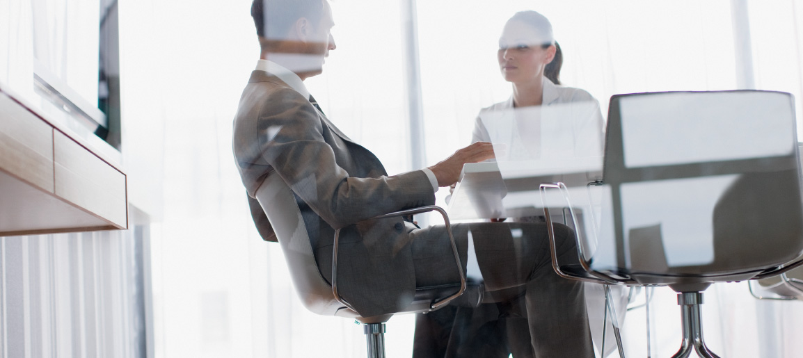 Two business professionals meeting in glass-walled office with laptop, discussing strategy in bright modern corporate conference room
