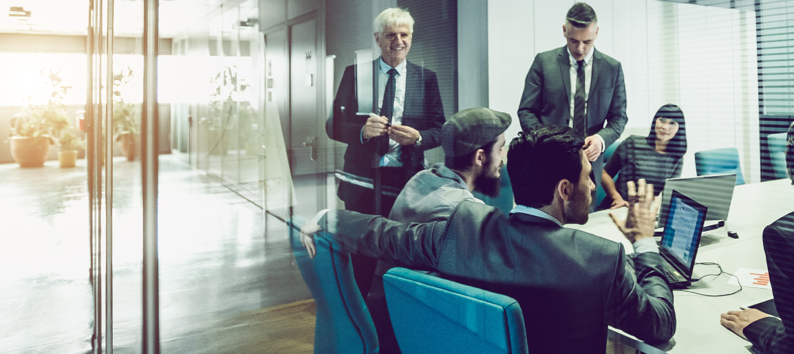 Business team meeting in glass-walled conference room with laptops and documents, colleagues discussing strategy in modern office setting