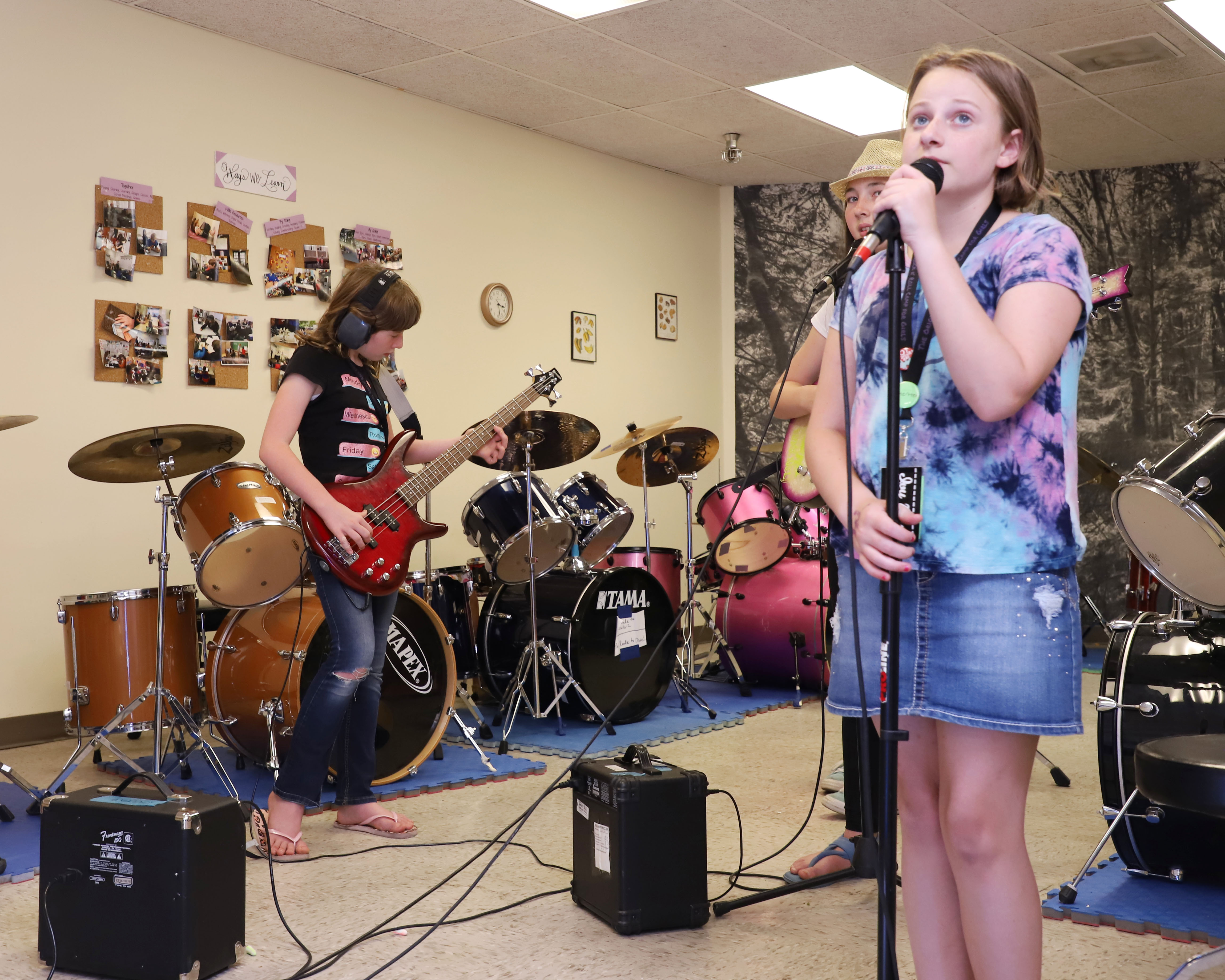 Image of a girl singing at a microphone and a girl in the background playing a guitar.