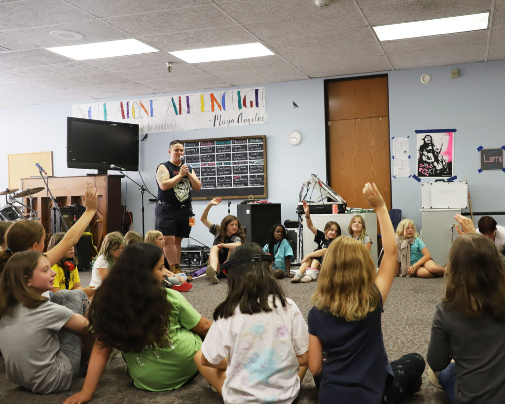 Image of a group of girls sitting in a circle with a teacher talking on a mic at Rock 'N Roll Camp for Girls.