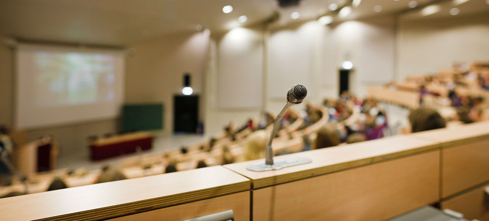 Microphone in lecture hall overlooking audience and presentation screen, university auditorium setup for public speaking and seminars