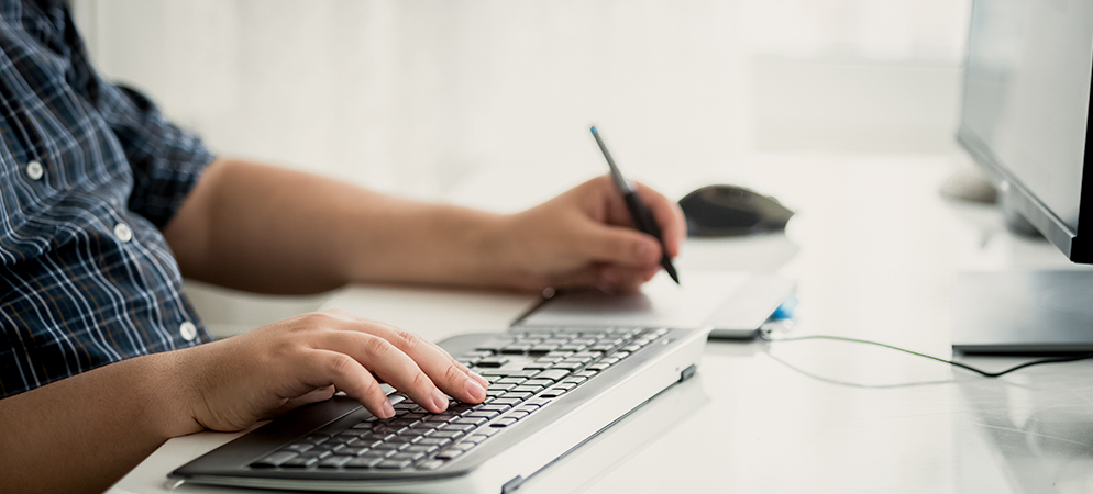 Person typing on keyboard and taking notes with pen at desk, using computer for work, study, and productivity in home office setup