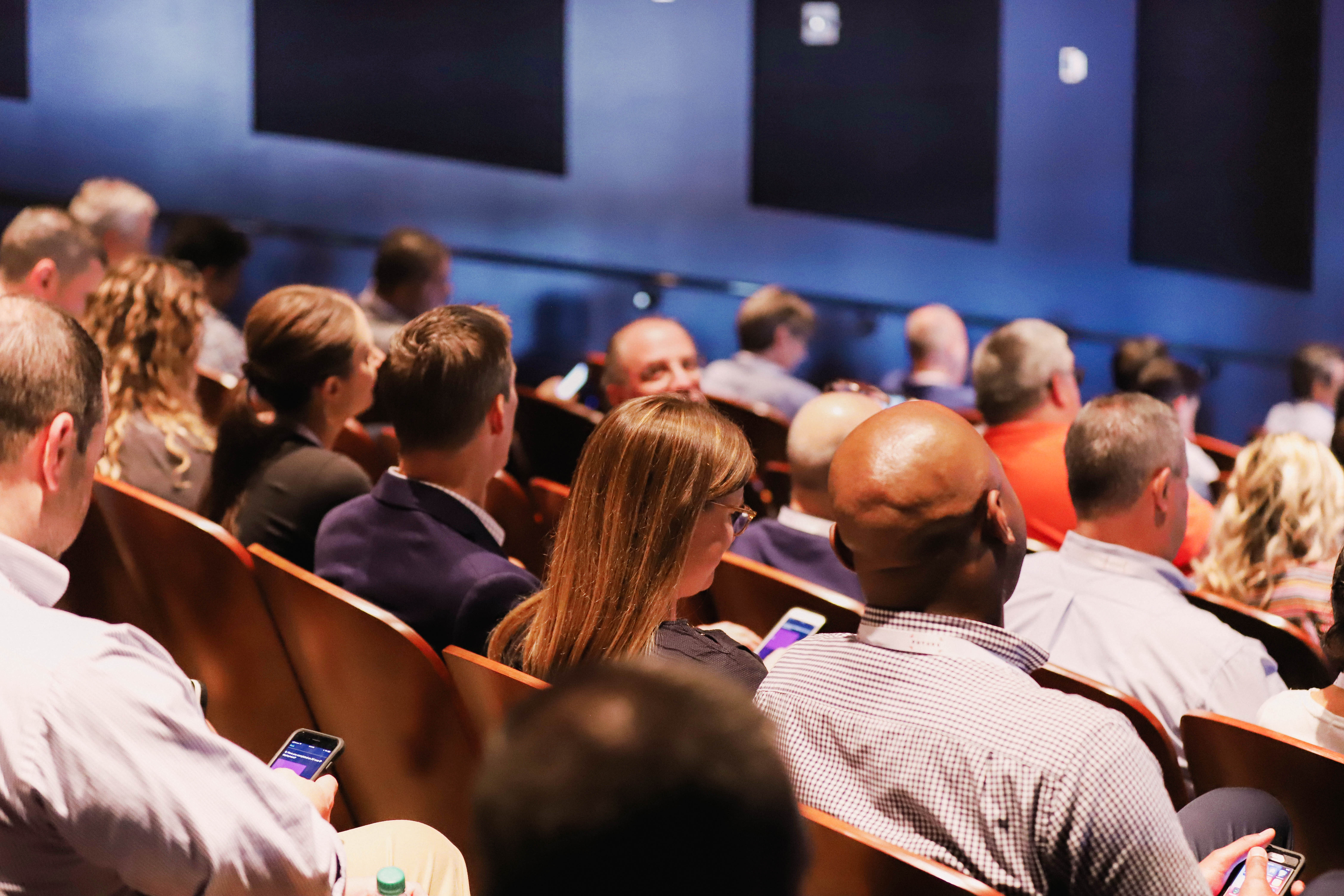 Audience seated in conference auditorium using smartphones, business event attendees waiting for keynote session in modern lecture theater.