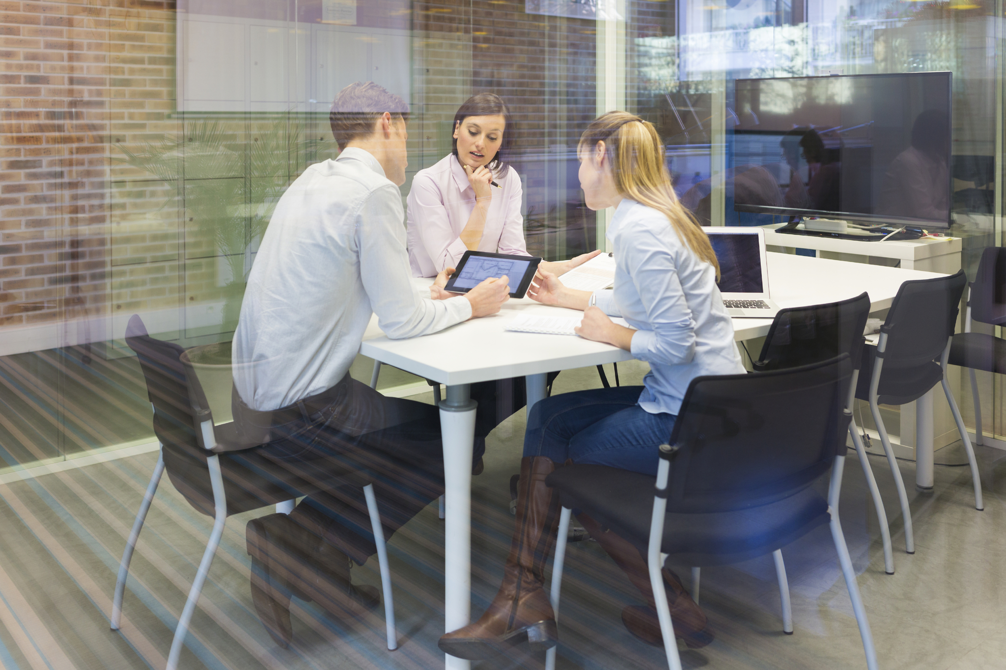 Business team meeting in glass conference room using tablet and laptop, collaborative project planning in modern office workspace.