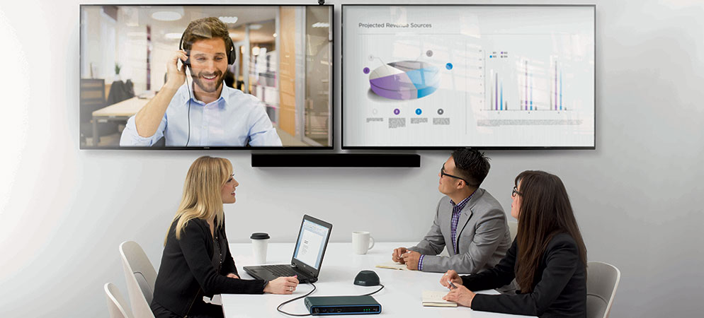 Business team in video conference meeting room watching remote presenter on dual displays with charts for hybrid collaboration and presentations
