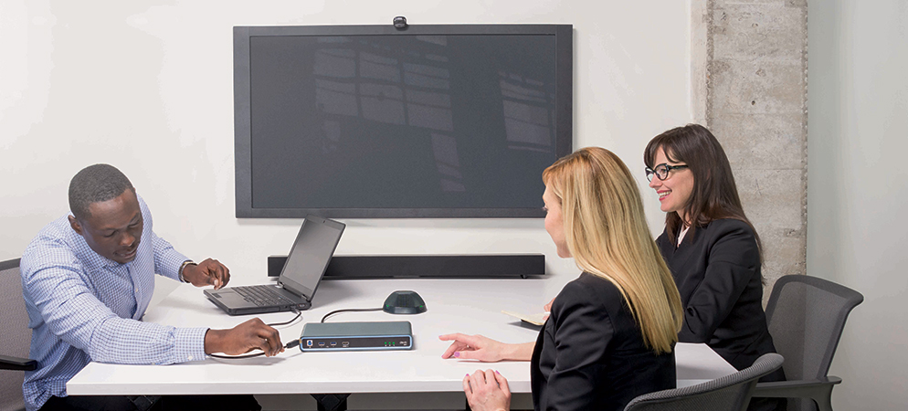 Modern conference room setup with team meeting, laptop and tabletop speakerphone, and large wall display for video conferencing and presentations
