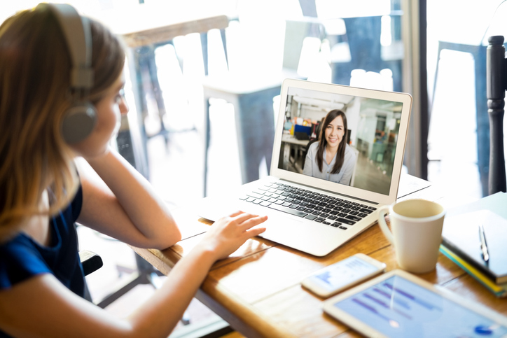 Woman wearing headset on video call with colleague on laptop, remote work online meeting at home office desk with coffee and tablet.