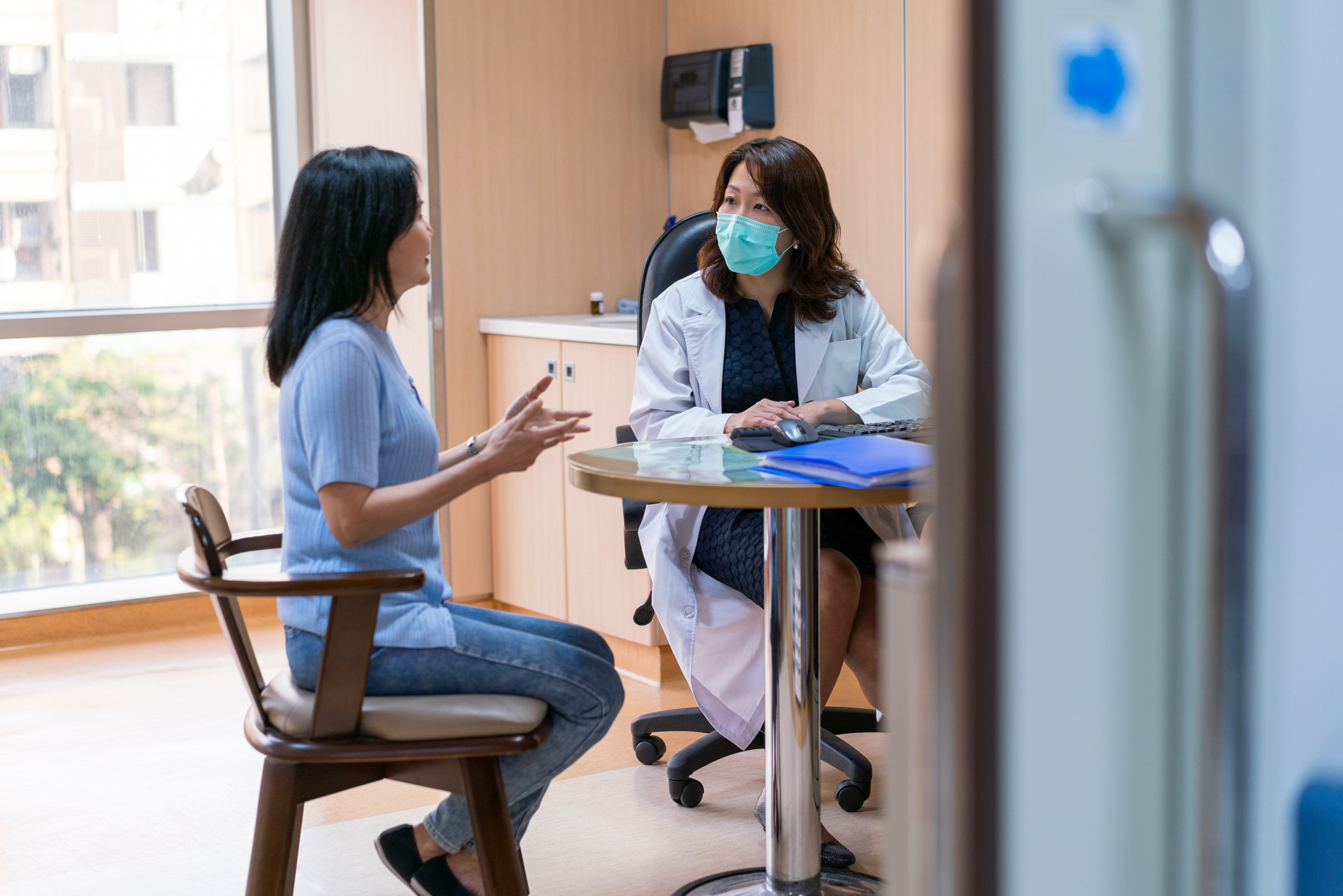 Doctor wearing face mask consulting with patient in medical office, healthcare appointment discussion and clinical care in hospital exam room.