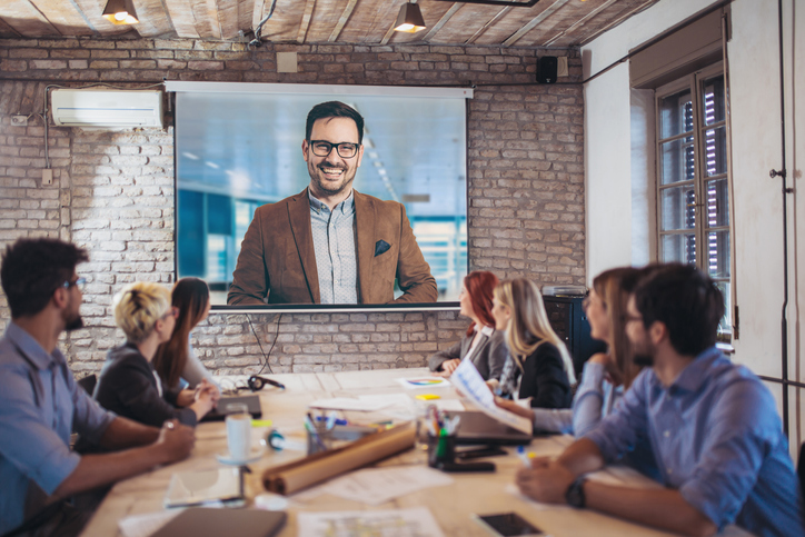 Team in conference room watching remote presenter on large screen, hybrid video meeting and business collaboration in modern office.