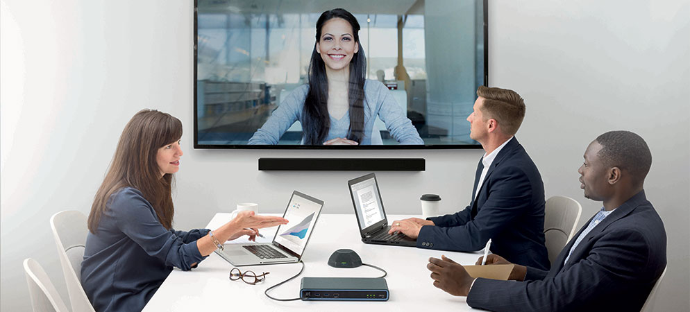 Team video conferencing in modern meeting room with laptops, tabletop speakerphone, and large display showing remote participant on screen