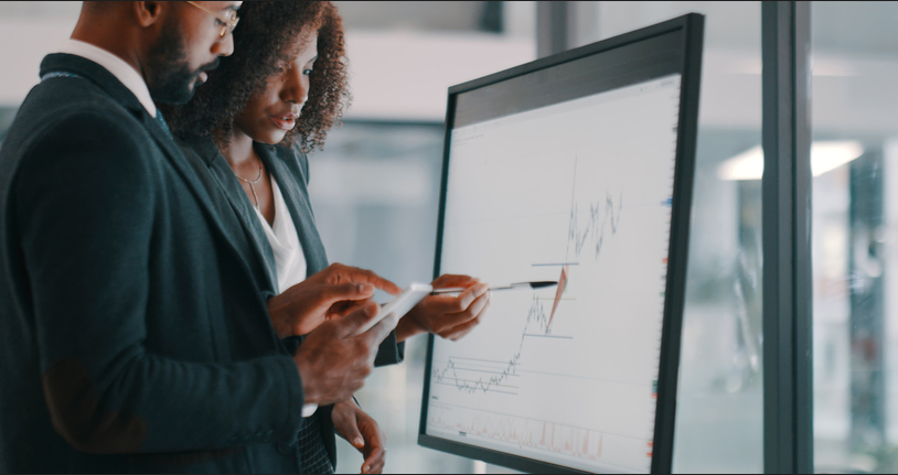 Business analysts reviewing financial charts on large monitor, teamwork with tablet and stylus, data analysis and investment strategy in office.
