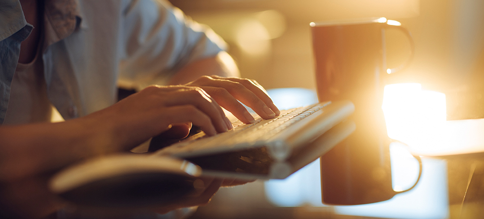 Close-up of hands typing on keyboard beside coffee mug in warm morning light, ideal for remote work and productivity theme
