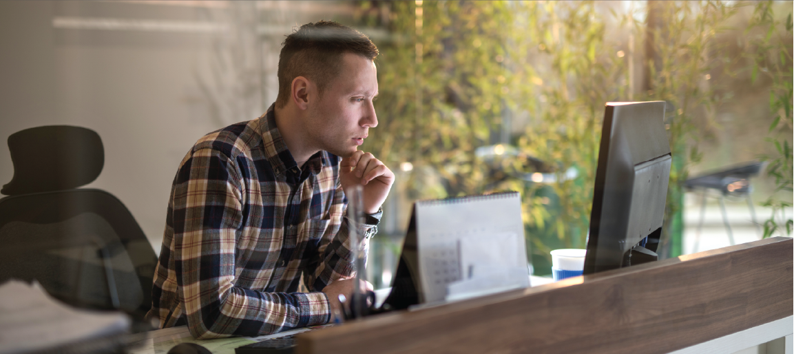 Focused professional working at desktop computer in modern office, employee analyzing data on monitor with natural light and plants.