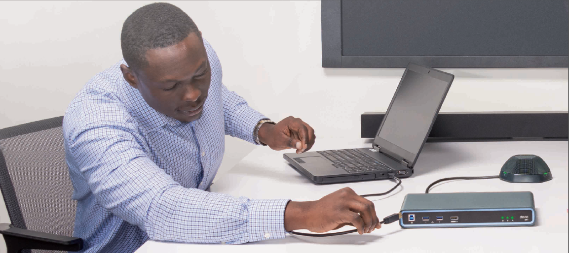 IT technician connecting Biamp Devio conferencing hub to laptop in meeting room, setting up USB audio and speakerphone for collaboration.