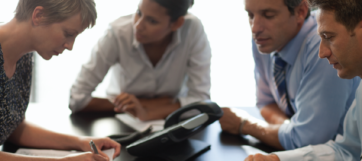 Business team collaborating at conference table with office phone, discussing project notes and strategy in corporate meeting room.