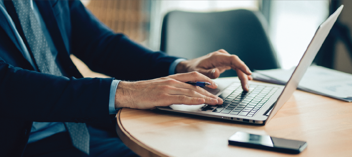 Businessman typing on laptop at desk with pen and smartphone, working on reports in modern office setting with productivity focus