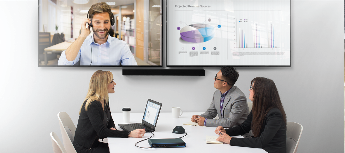 Hybrid video conference meeting with remote presenter on dual screens, team using laptop and Biamp conferencing hub for clear audio collaboration.