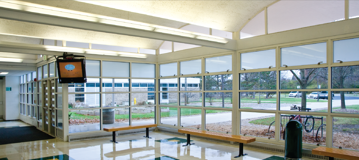 Bright school hallway entrance with large windows, benches, and wall-mounted TV display, showcasing modern campus lobby and waiting area