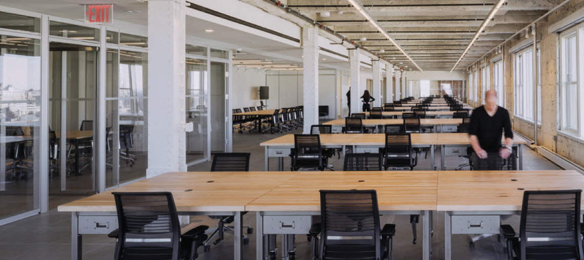 Modern open-plan office with long shared desks and glass meeting rooms, coworking workspace interior with rows of workstations and natural light.