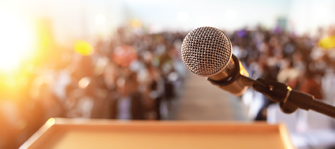 Close-up microphone on podium facing large audience, public speaking and conference presentation stage, event keynote and speech setup.