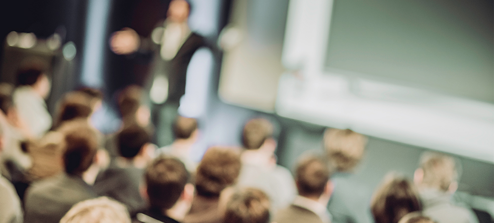 Blurred audience watching speaker presentation in conference hall, business seminar or training event with projector screen and crowd seating