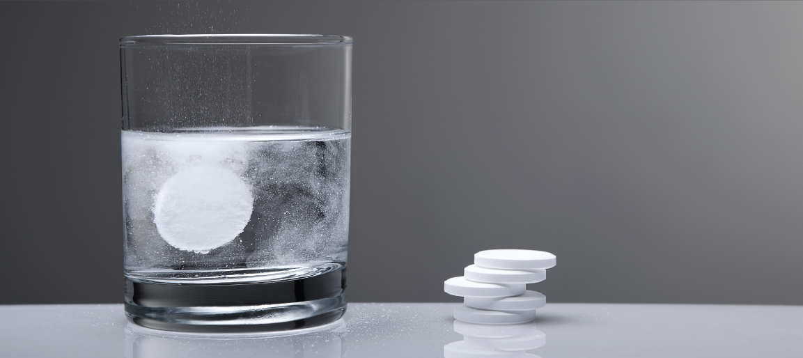 Effervescent tablet dissolving in glass of water with fizz bubbles, next to stacked tablets on table, healthcare and hydration concept