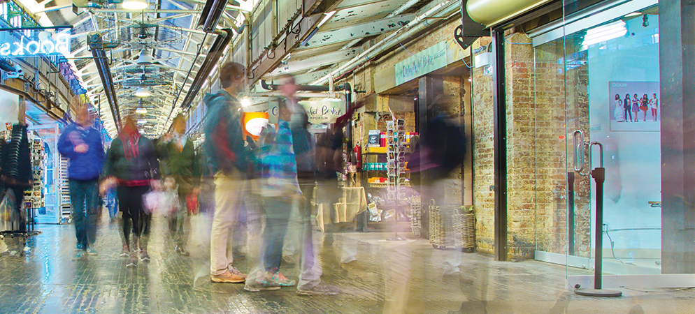 Blurred shoppers walking through vibrant indoor market corridor with exposed brick, shops, and bright retail lighting