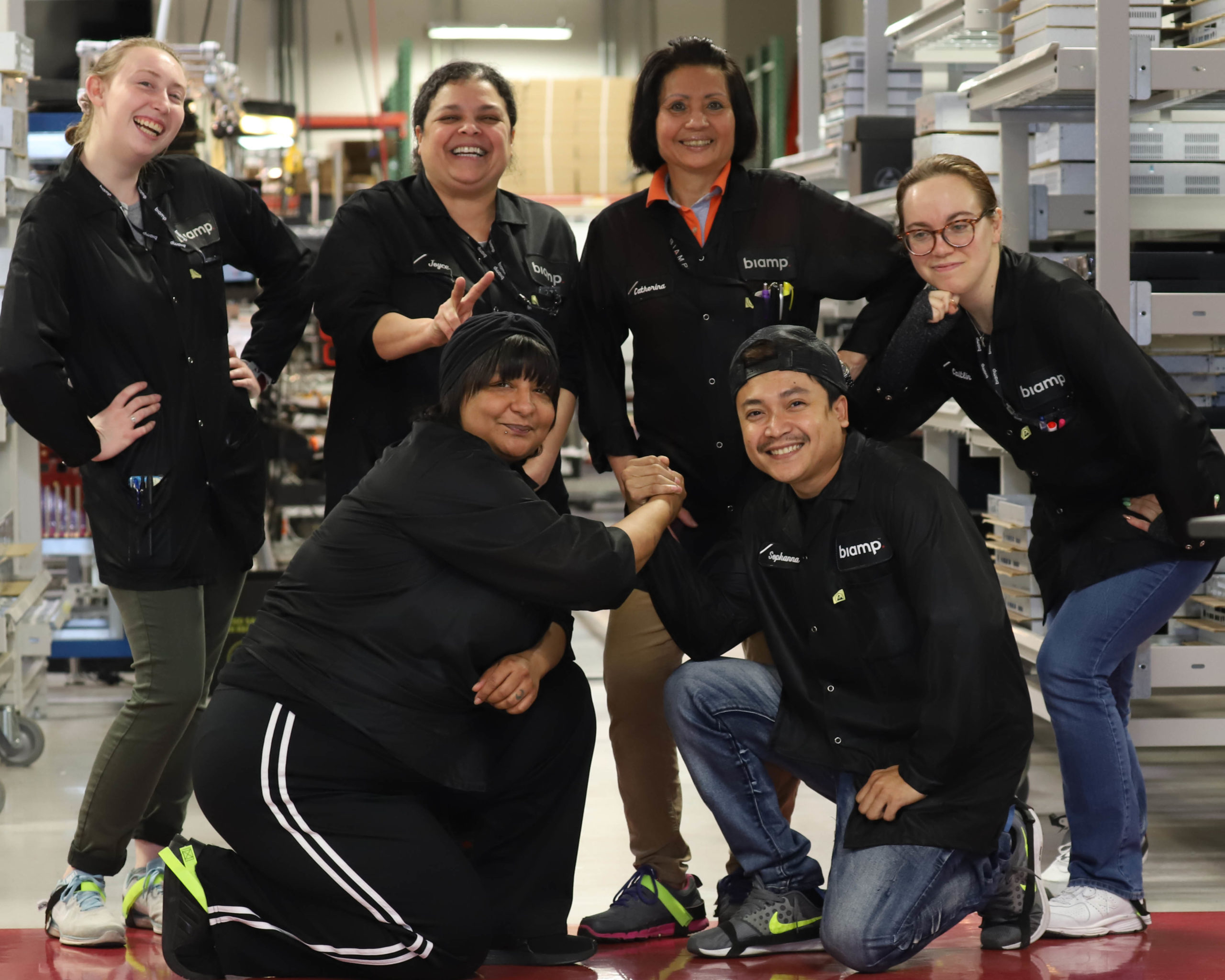 Biamp manufacturing team group photo in factory, employees in branded work shirts celebrating teamwork on production floor with equipment.