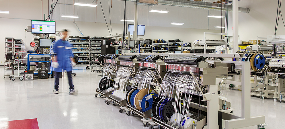 Electronics manufacturing floor with assembly equipment, cable reels, and technician walking through modern production facility