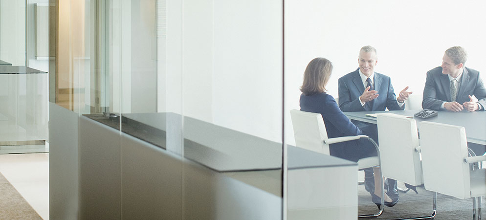 Business meeting in bright glass-walled conference room with three professionals discussing at table in modern corporate office