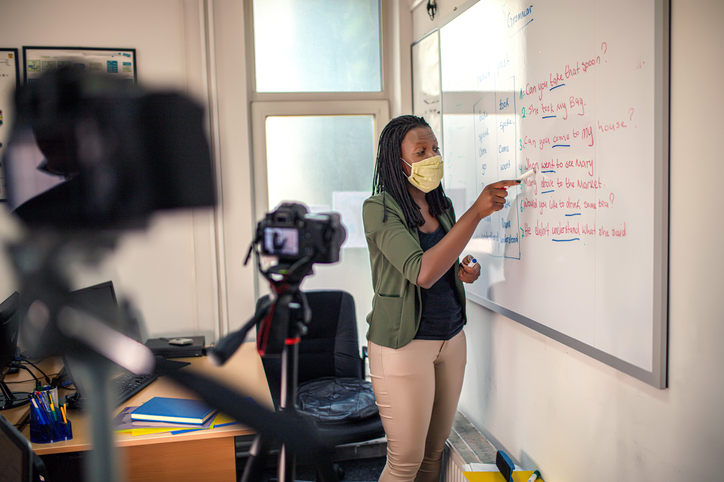 Teacher wearing face mask recording online lesson on whiteboard with camera, remote learning classroom setup and virtual education training.