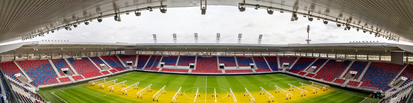 Wide panoramic view of empty soccer stadium with red and blue seats, green pitch and roof canopy, modern sports arena architecture.