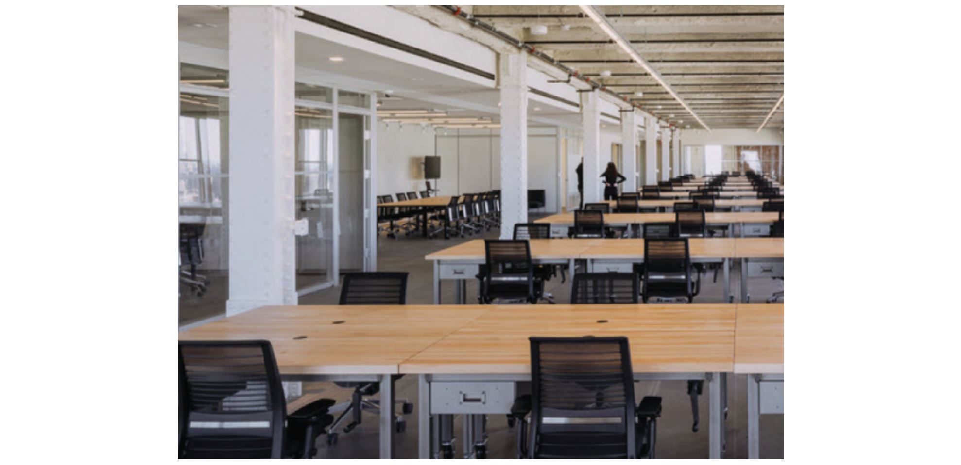 Modern open office workspace with long shared desks, rolling chairs, and glass meeting rooms in large corporate floor plan