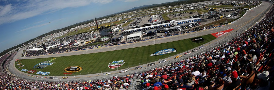 Wide aerial view of NASCAR race track with packed grandstands and cars on oval circuit, motorsport event crowd and stadium atmosphere.