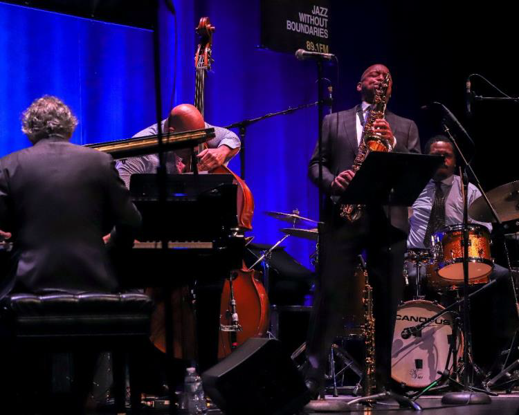 Live jazz quartet on stage with saxophone, piano, bass and drums under blue lighting, concert performance at jazz club or festival.