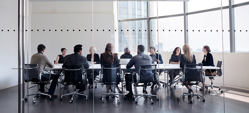 Large team meeting in glass-walled conference room with city views, people seated around long table for corporate collaboration and planning