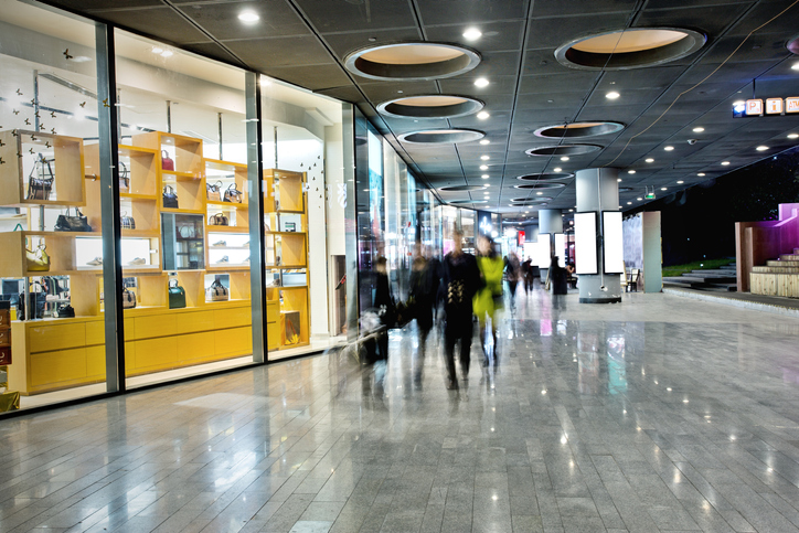 Modern shopping mall corridor with glass storefronts and blurred shoppers, bright retail interior walkway, urban commercial space and foot traffic.