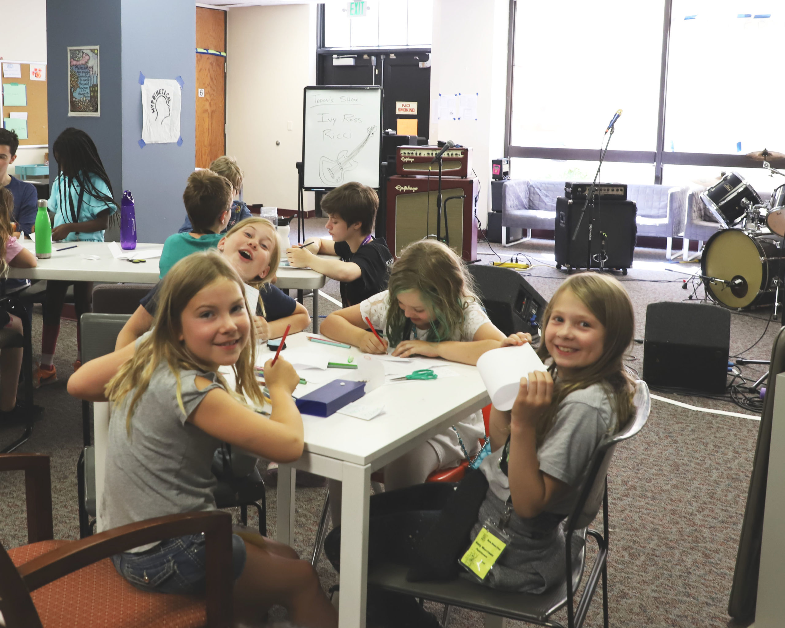 Kids music camp classroom with students drawing at tables, band rehearsal gear in background, creative arts activity and youth music education.