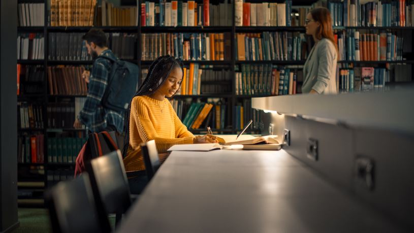 Student studying at library desk with bookshelves in background, focused reading and note-taking in quiet academic space