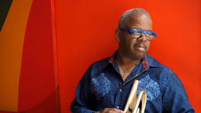 Jazz trumpeter Terence Blanchard holding trumpet against bold red wall, portrait of performer with blue glasses, live music and concert artist promotional photo.