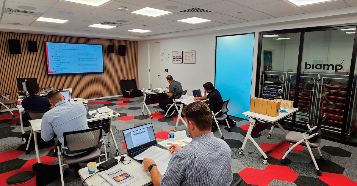 Biamp training classroom with students working on laptops, presentation screen, and visible AV rack room for hands-on audio video education
