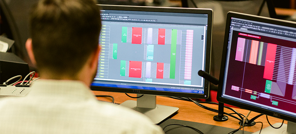 Engineer monitoring dual screens with AV system configuration software and signal routing diagrams in control room or IT operations workspace