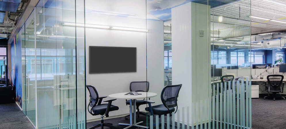 Glass-walled meeting room in modern open-plan office with round table, task chairs, and wall-mounted display for video conferencing